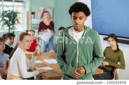 Sad African-American boy stands in study room, group of classmates behind him. Sad African-American boy stands in study room, group of classmates behind him. 138542959