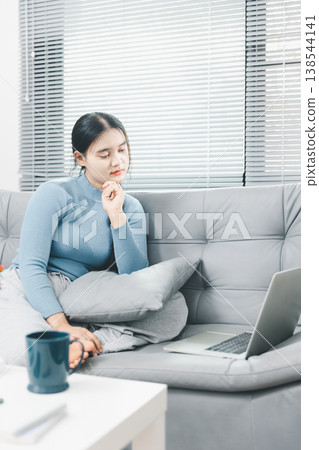 Asian woman sitting on sofa with pillow and looking at laptop at home, showing remote work, online learning, focused browsing and cozy digital lifestyle in bright modern interior. 138544141