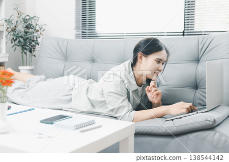 Asian woman using laptop while lying on sofa at home, showing relaxed remote work, online browsing and cozy digital lifestyle in a bright modern living room with casual comfort. 138544142
