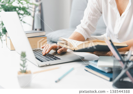 Hands studying at home, reading open book while typing on laptop on white desk with stacked books and stationery in bright daylight. Online learning and exam preparation concept. education, learning 138544388