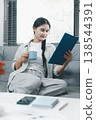 Asian woman reviewing clipboard with coffee and laptop on sofa at home, showing remote work, document planning, home office routine and calm productivity in a bright modern living room. 138544391