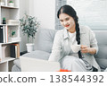 Asian woman using laptop while holding coffee cup on sofa at home, showing remote work, online browsing, digital productivity and cozy home office lifestyle in bright interior. 138544392