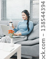 Asian woman using laptop on sofa at home while holding takeaway coffee cup, showing remote work, online productivity and cozy digital lifestyle in bright modern living room with copy space. 138544394