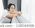 Sad woman leaning on sofa by bright window at home, feeling depressed and anxious. Mental health, loneliness and burnout concept for therapy, counseling, support and awareness campaigns, copy space. 138544506