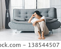 Depressed woman sitting on floor beside gray sofa, leaning head on hand and staring ahead in bright living room. Mental health anxiety, loneliness, burnout concept with copy space. depression 138544507