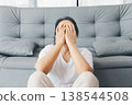 Depressed woman sitting on floor in front of gray sofa, covering face with hands at home. Mental health anxiety, stress, burnout and loneliness concept for therapy and awareness campaigns. stress 138544508