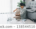 Woman working from home on laptop, reviewing printed charts and taking notes on the floor beside sofa with coffee and smartphone. Remote work, home office productivity concept for telecommuting. 138544510