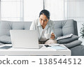 Focused woman working from home on laptop at coffee table, holding coffee cup with phone and notebook on table in bright living room. Remote work, telecommuting, home office productivity concept. 138544512