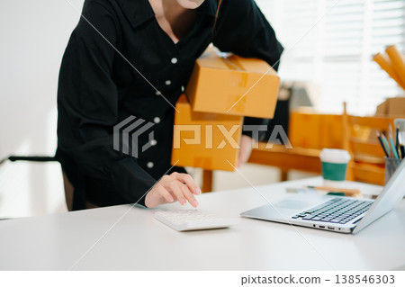 Entrepreneurial Asian woman prepares parcels and checks online orders on tablet and laptop for delivery online shopping and small business success in office 138546303