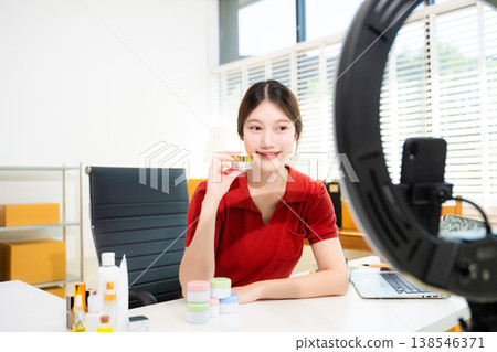 Smiling Asian woman influencer holding cream jar and live streaming for online business in her home office studio 138546371