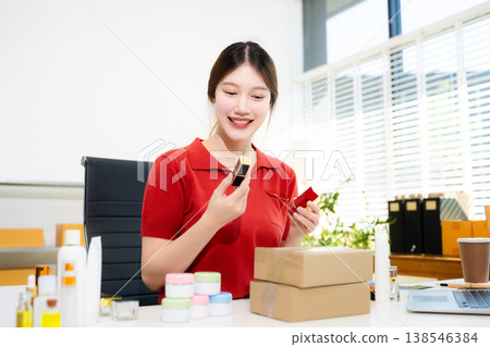 Social media influencer marketing. Smiling Asian beauty vlogger reviewing lipstick and cosmetic products while filming with a ring light in her home office. Social media influencer marketing. Smiling Asian beauty vlogger reviewing lipstick and cosmetic products while filming with a ring light in her home office. 138546384