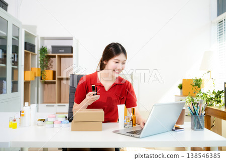 Social media influencer marketing. Smiling Asian beauty vlogger reviewing lipstick and cosmetic products while filming with a ring light in her home office. Social media influencer marketing. Smiling Asian beauty vlogger reviewing lipstick and cosmetic products while filming with a ring light in her home office. 138546385