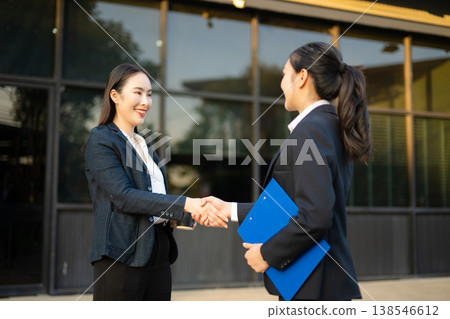 Two smiling Asian businesswomen in suits shaking hands while standing outside office building with coffee and clipboard Two smiling Asian businesswomen in suits shaking hands while standing outside office building with coffee and clipboard 138546612