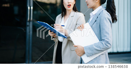 Two business women discussing documents and clipboard outside modern office, professional teamwork, corporate communication, management, agreement Two business women discussing documents and clipboard outside modern office, professional teamwork, corporate communication, management, agreement 138546638