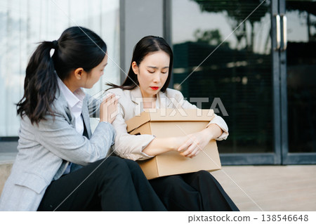 Stressed businesswoman holding a box after job loss, supported by colleague. Workplace stress, unemployment, burnout, mental health, corporate crisis and support concept. 138546648