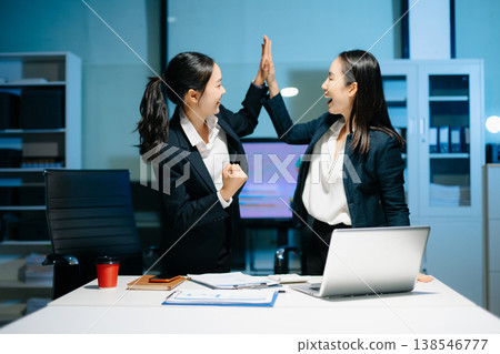 Two businesswomen celebrating success after achieving goals at office desk. Teamwork, motivation, leadership, startup growth and modern corporate workplace. 138546777