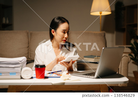 Stressed Asian woman using calculator and laptop for financial accounting and tax report late at night at home. Stressed Asian woman using calculator and laptop for financial accounting and tax report late at night at home. 138546829