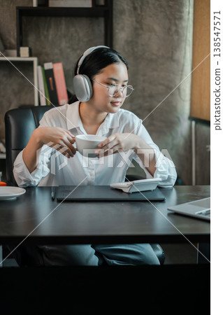 Woman in office wearing headphones, holding coffee, and using a tablet. Modern workspace with relaxed atmosphere. Woman in office wearing headphones, holding coffee, and using a tablet. Modern workspace with relaxed atmosphere. 138547571