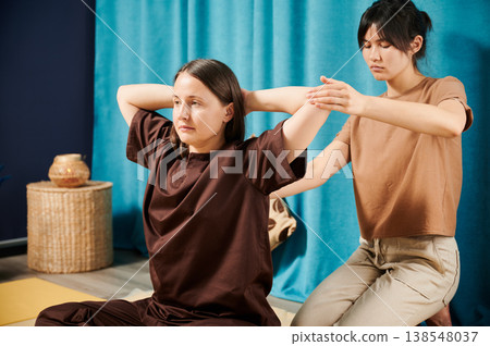 Masseuse making thai yoga massage. Woman therapist doing traditional massage treatment for female patient indoors. Client sitting on mat, enjoying hand massage in calm atmosphere. Masseuse making thai yoga massage. Woman therapist doing traditional massage treatment for female patient indoors. Client sitting on mat, enjoying hand massage in calm atmosphere. 138548037