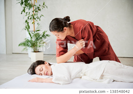 Masseuse making thai yoga massage. Woman therapist doing traditional massage treatment for female patient using elbow. Client lying on mat, meditating, enjoying hand massage in calm atmosphere. Masseuse making thai yoga massage. Woman therapist doing traditional massage treatment for female patient using elbow. Client lying on mat, meditating, enjoying hand massage in calm atmosphere. 138548042