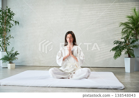 Beautiful woman in white dress sitting on mat in lotus position before Thai massage, practicing yoga, showing namaste sign. Beautiful woman in white dress sitting on mat in lotus position before Thai massage, practicing yoga, showing namaste sign. 138548043
