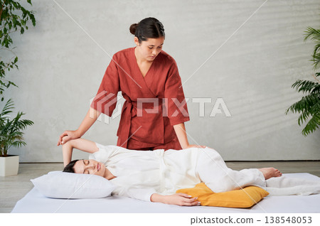 Masseuse making thai yoga massage. Woman therapist doing traditional massage treatment, stretching female patient's body. Client lying on mat, enjoying hand massage in calm atmosphere. 138548053