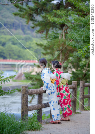 The backs of two women in yukata (summer kimonos) leaning against a wooden railing over the Uji River, gazing at the Asagiri Bridge. Uji City, Kyoto Prefecture, Japan. 138548396