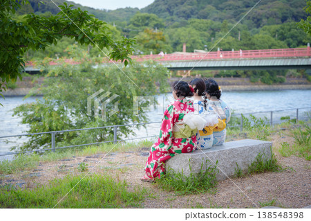 The backs of three women in yukata (summer kimonos) sitting on a stone bench by the Uji River, gazing at the Asagiri Bridge. Uji City, Kyoto Prefecture, Japan. The backs of three women in yukata (summer kimonos) sitting on a stone bench by the Uji River, gazing at the Asagiri Bridge. Uji City, Kyoto Prefecture, Japan. 138548398