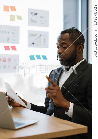 An African American man wearing a suit and headset gestures while holding a document, looking towards a laptop screen in a modern office setting with charts on the wall An African American man wearing a suit and headset gestures while holding a document, looking towards a laptop screen in a modern office setting with charts on the wall 138549028