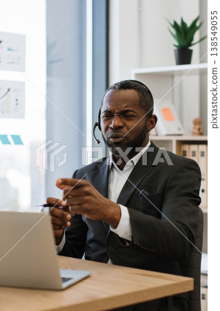 An African American man wearing a headset and suit expresses frustration while gesturing during a video conference call on his laptop An African American man wearing a headset and suit expresses frustration while gesturing during a video conference call on his laptop 138549055
