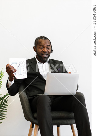 An African American man in a suit wearing a headset presents a financial growth chart while on a video call with his laptop An African American man in a suit wearing a headset presents a financial growth chart while on a video call with his laptop 138549063