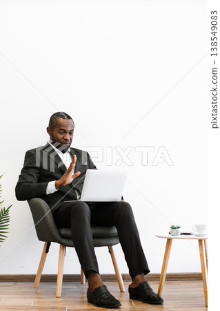 An African American man in a suit sits in a chair, holding a laptop and gesturing during a video conference call 138549083