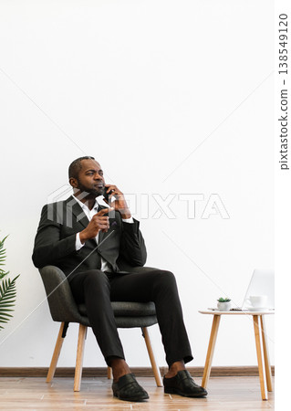 An African American man in a suit sits in a chair, talking on his mobile phone and gesturing with his hand, with a laptop on a nearby table An African American man in a suit sits in a chair, talking on his mobile phone and gesturing with his hand, with a laptop on a nearby table 138549120