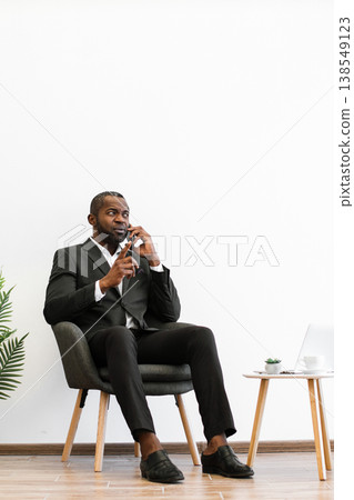 An African American man in a suit sits in a chair while talking on his mobile phone, looking concerned An African American man in a suit sits in a chair while talking on his mobile phone, looking concerned 138549123