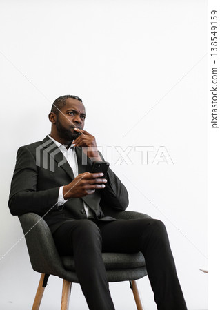 An African American man in a suit sits in a chair, holding a smartphone and contemplating with his finger on his chin An African American man in a suit sits in a chair, holding a smartphone and contemplating with his finger on his chin 138549159