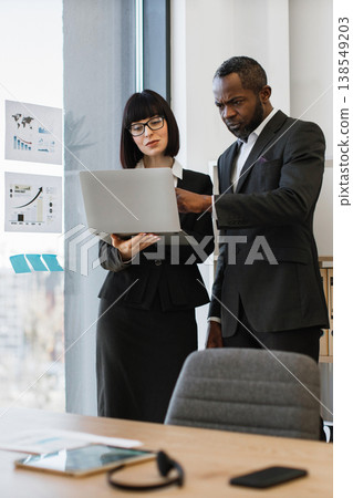 A focused African American man and a woman in business attire collaborate over a laptop, reviewing charts and data on a glass wall A focused African American man and a woman in business attire collaborate over a laptop, reviewing charts and data on a glass wall 138549203