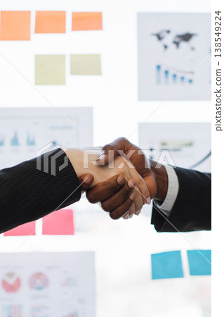 Two professionals, one African American man and one woman, shake hands in a gesture of agreement and partnership in a modern office setting with charts and sticky notes visible Two professionals, one African American man and one woman, shake hands in a gesture of agreement and partnership in a modern office setting with charts and sticky notes visible 138549224