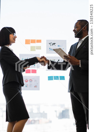 Two professionals, an African American man and a woman, shake hands in front of a glass wall with charts and sticky notes, symbolizing a successful deal 138549225