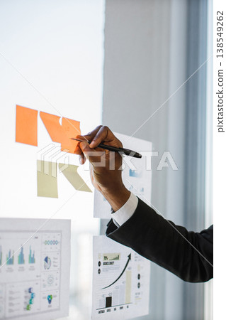 An African American hand in a suit writes on sticky notes attached to a glass wall with charts and graphs in the background An African American hand in a suit writes on sticky notes attached to a glass wall with charts and graphs in the background 138549262