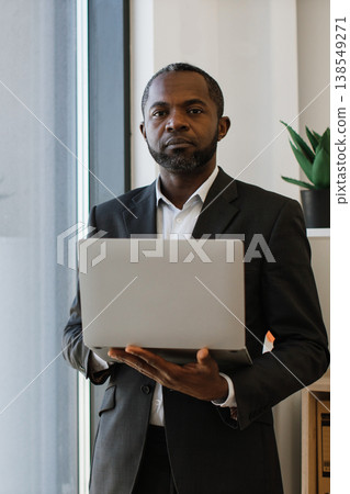 A professional African American man in a suit holds a laptop computer while standing near a window in an office environment 138549271