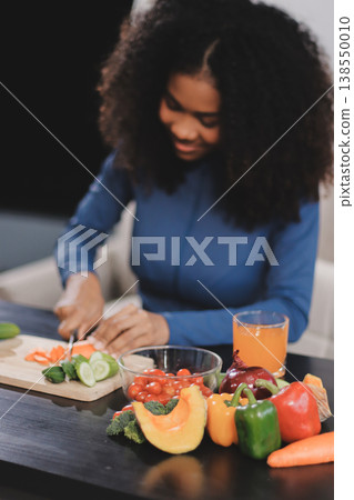 Young nutritionist showing bell peppers during an online video call consulting about healthy eating, sitting at home at a table with vegetables, laptop and measuring tape 138550010