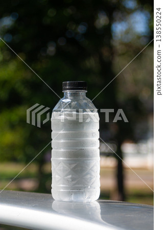 A cold plastic water bottle with condensation stands outdoors, catching sunlight against green blur. A cold plastic water bottle with condensation stands outdoors, catching sunlight against green blur. 138550224