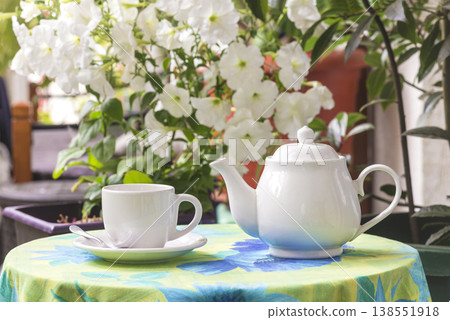 White cup of tea, tea pot on a table covered by tablecloth against white blooming flowers 138551918