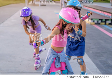 A group of children in sports helmets and protective gear ride roller skates and skateboards in a city skate park A group of children in sports helmets and protective gear ride roller skates and skateboards in a city skate park 138553136