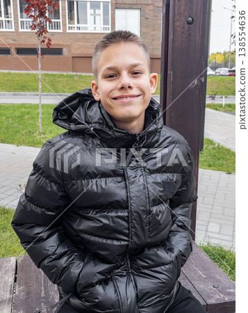 Teen joy on street. Energetic teenager with bright smile relaxing on city park bench in stylish jacket Teen joy on street. Energetic teenager with bright smile relaxing on city park bench in stylish jacket 138554636