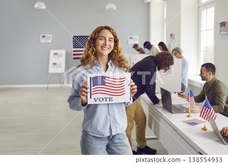 Happy young woman standing at polling station and holding VOTE poster with USA flag Happy young woman standing at polling station and holding VOTE poster with USA flag 138554933