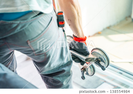 Worker carrying window glass panel using suction cup lifter during installation or replacement in apartment. Safe handling, professional equipment and glass transport process concept. Worker carrying window glass panel using suction cup lifter during installation or replacement in apartment. Safe handling, professional equipment and glass transport process concept. 138554984