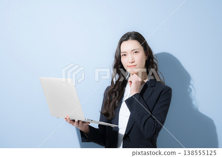 A cool, business-suited woman with her hand on her chin, holding a laptop, is shown against a light blue background, in a thoughtful, contemplative pose. (Image caption: Job hunting) 138555125