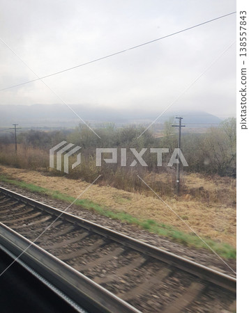View of railway tracks and foggy mountains from train window. Gloomy spring landscape, metal rails, telegraph poles View of railway tracks and foggy mountains from train window. Gloomy spring landscape, metal rails, telegraph poles 138557843