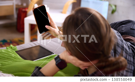 Woman lying on sofa using smartphone with green dress and tablet nearby 138558049